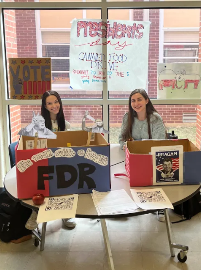 Michaela Scott and Charlotte Penberthy sit behind a table outside of the cafeteria, supervising the canned food drive and voter registration signups. Handmade posters are displayed behind them, further advertising the event.
